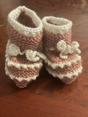 Rosa och vita stickade babytofflor - Supersöta ny  handstickade babytofflor i rosa och vitt med dekorativa virkade kanter och små rosetter framtill. Tofflorna har en mjuk och mysig känsla, perfekta för små fötter. Passar dig som gillar gulliga detaljer och handgjorda accessoarer.