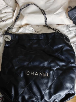Svart  quiltad Hobo väska - Snygg svart quiltad handväska med silverfärgad kedjerem och logotyp framtill. Väskan är tillverkad i mjukt läder och har en rund metallbricka med gravyr. Materialet ska vara läder enligt butiken där jag köpte. Kan dock ej garantera det till 100% men jag tycker det känns och doftar som läder Perfekt för dig som vill ha en klassisk och trendig accessoar. Rimlig och får plats med mycket 