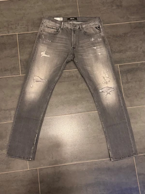 Grå slitna jeans från Replay - Säljer ett par grå jeans från Replay med slitningar och distressed detaljer på framsidan. Jeansen har fem fickor, raka ben och en snygg tvättad look. Perfekta för en avslappnad och trendig stil.