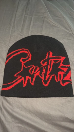 Cortez mössa - Säljer en svart mössa med coolt rött graffiti-inspirerat mönster. Mössan är stickad och har en mjuk, stretchig passform som sitter skönt på huvudet. Perfekt för dig som gillar streetwear och vill sticka ut med en unik accessoar.