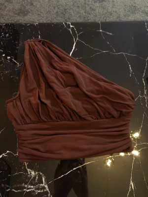 Två toppar i brunt och vitt - Säljer två snygga, axelbandslösa toppar i brunt och vitt. Båda har en one shoulder-design med draperad, rynkad look och bred linning nedtill. Perfekta för att sticka ut och matcha med höga jeans eller kjol. Materialet känns mjukt o lyxigt. En för 30 eller båda för 50