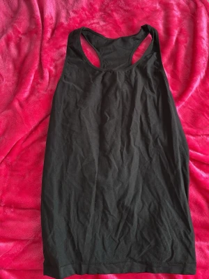 Svart ärmlös linnetopp - En enkel och stilren svart linnetopp med brottarrygg. Tillverkad i ett stretchigt material av 90% polyamid och 10% elastan, vilket gör den bekväm och följsam. Perfekt att matcha med jeans eller shorts för en avslappnad look. Fick den av gammelmormor och har aldrig använt!