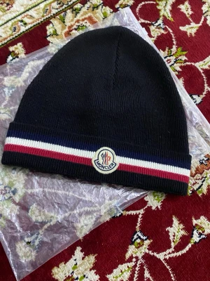 Svart Moncler mössa med ränder - Snygg svart mössa från Moncler med klassiska ränder i rött, vitt och blått längs kanten. Mössan har Moncler-logga framtill och är stickad i mjukt material som håller dig varm. Perfekt accessoar för att lyfta din stil under kyliga dagar.