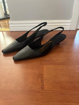 Svarta slingback pumps i skinn - Stilrena svarta pumps med låg klack och slingback-rem. Skorna har spetsig tå och är tillverkade i slätt skinn som ger en elegant look. Perfekta för dig som gillar minimalistisk och klassisk stil.