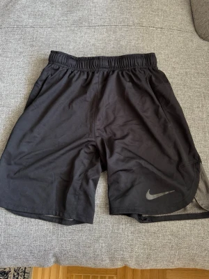 Svarta träningsshorts från Nike - Säljer ett par svarta träningsshorts från Nike med elastisk midja och diskret Nike-logga på vänster ben. Shortsen har en loose passform och är tillverkade i ett lätt och snabbtorkande syntetmaterial, perfekta för gymmet eller löprundan.