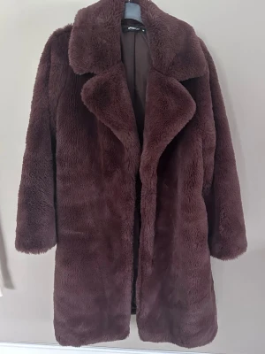 Brun fuskpäls kappa från Gina Tricot - Mysig brun fuskpäls kappa från Gina Tricot med bred krage och rak passform. Kappan har långa ärmar och är helfodrad för extra komfort. Perfekt för att hålla värmen under kyliga dagar och ger en trendig look.