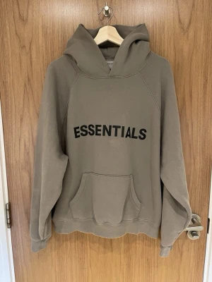 Brun/Mörkgrön Essentials hoodie med huva - Säljer en brun/mörkgrön hoodie från Essentials med stor svart logga på bröstet. Hoodien har en rymlig huva och en klassisk magficka framtill. Tillverkad i mjukt bomullsmaterial och har ribbade muddar vid ärmslut och nederkant. Perfekt för en chill och avslappnad stil.