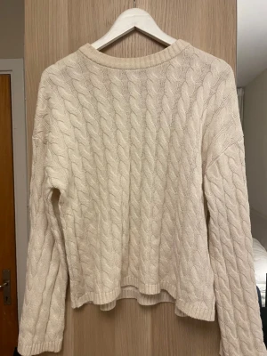 Beige kabelstickad tröja Gina Tricot - Kabelstickad beige tröja från Gina Tricot med rund halsringning och långa ärmar. Tröjan har ett klassiskt flätmönster och ribbstickade muddar vid hals, ärmslut och nederkant. Perfekt för lager-på-lager och enkel att matcha.