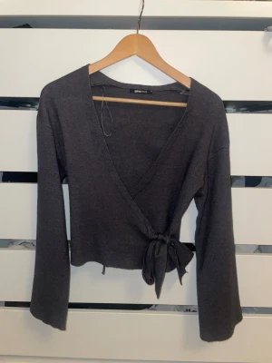 Mörkgrå omlottopp från Gina Tricot - Säljer en mörkgrå omlottopp från Gina Tricot med långa ärmar och knytband i midjan. Tröjan har en djup V-ringning och är tillverkad i mjukt stickat material. Perfekt för en trendig och avslappnad look.