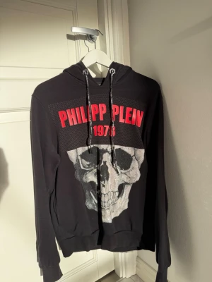 Äkta Philipp Plein hoodie - Svart hoodie från Philipp Plein med stor dödskalle i swarowski stenar på framsidan och röd text 'PHILIPP PLEIN 1978'. Huva med snören och ribbade muddar. Perfekt för dig som gillar ovanliga plagg och som vill sticka ut med. Nypris 10 000 kr +.