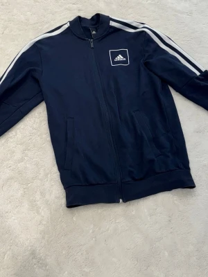 Mörkblå Adidas ziptröja med ränder - Mörkblå ziptröja från Adidas med vita ränder längs ärmarna och Adidas-logga på bröstet. Tröjan har två fickor framtill och ribbade muddar. Perfekt för en sportig och avslappnad stil.