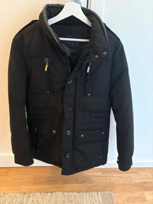 Svart pufferjacka med huva ifrån Zara - Snygg svart pufferjacka med avtagbar huva och flera fickor med dragkedja och knappar. Jackan har hög krage, axelklaffar och quiltad design som håller värmen. Perfekt för kalla vinterdagar och har en modern, stilren look.