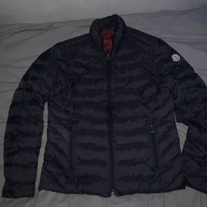 Svart Moncler pufferjacka - Säljer en svart pufferjacka från Moncler med klassisk quiltad design och hög krage. Jackan har två dragkedjeförsedda fickor framtill och ikonisk Moncler-logga på ärmen. Materialet är glansigt och dunfyllt för extra värme under kalla dagar.