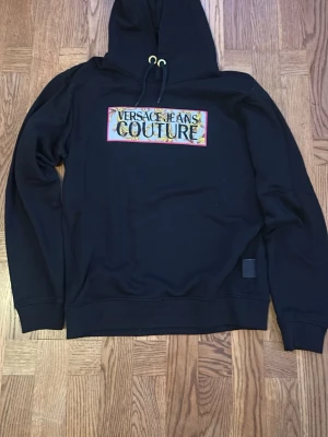 Svart hoodie Versace Jeans Couture - Snygg svart hoodie från Versace Jeans Couture med färgglad broderad logga på bröstet. Klassisk huva med snörning och guldfärgade öljetter. Mjuk bomullskänsla och relaxed fit, perfekt för en trendig streetwear-look.