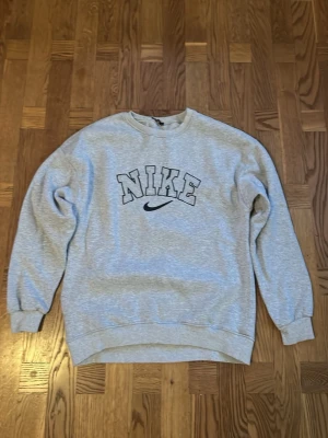 Grå sweatshirt från Nike - Säljer en klassisk grå sweatshirt från Nike med stor logga och swoosh broderat på bröstet. Tröjan har rund halsringning, ribbade muddar vid ärmslut och nederkant samt en avslappnad passform. Tröjan är M men skulle säga att den mer passar S.