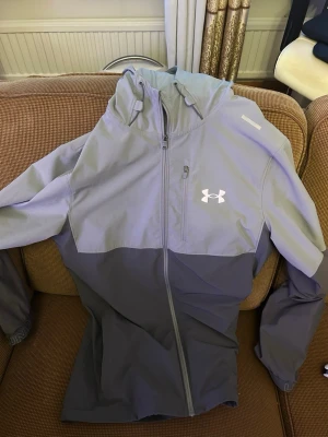 Grå vindjacka från Under Armour - Snygg tvåtonad grå vindjacka från Under Armour med huva och hel dragkedja framtill. Jackan har logga på bröstet och är tillverkad i lätt, vindtätt material. Perfekt för blåsiga dagar och sportiga outfits.