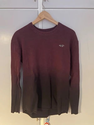 stickad tröja Hollister - Stickad tröja från Hollister i burgundy med snygg ombre-effekt som går över till svart nedtill. Tröjan har rund halsringning, ribbade muddar och Hollisters klassiska logga broderad på bröstet. Perfekt för dig som gillar stilrena detaljer och skön passform.