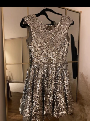 Glittrig silverklänning med paljetter - Superfin kort klänning i silver med massor av paljetter som glittrar. Klänningen har ärmlös design, rund halsringning och en öppen rygg. Perfekt för dig som vill sticka ut och glänsa. Materialet är syntet och klänningen har en figurnära överdel med utställd kjol.