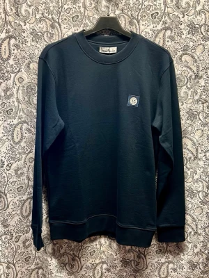 Mörkblå sweatshirt från Stone Island - Säljer en stilren mörkblå sweatshirt från Stone Island med klassisk rund halsringning och lång ärm. Tröjan har en liten patch med logga på bröstet och ribbade muddar vid ärmslut och nederkant. Perfekt för dig som gillar streetwear och enkel design. Finns i flera storlekar. Kontakta oss i samband med order. 