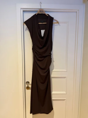 Brun draperad klänninh - Snygg brun klänning med draperad effekt och djup ringning fram. Klänningen har ett elegant fall och är ärmlös med breda axelpartier. Tillverkad i ett mjukt, stretchigt tyg som ger en smickrande siluett. Storlek S. Klänningen är helt ny. 
