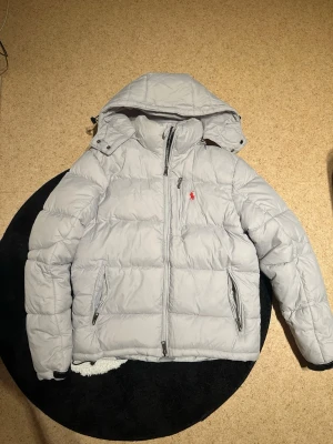 Ljusgrå dunjacka från Polo Ralph Lauren - Snygg ljusgrå dunjacka från Polo Ralph Lauren med avtagbar huva och dragkedja framtill. Jackan har två snedställda fickor med dragkedja och en bröstficka. Klassisk röd logga broderad på bröstet. Perfekt för kalla vinterdagar.