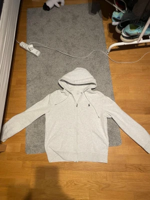 Ljusgrå hoodie från Ralph Lauren - Snygg ljusgrå hoodie med dragkedja från Ralph Lauren. Klassisk design med liten broderad logga på bröstet, två fickor fram och justerbar huva med snören. Perfekt för en chill och avslappnad stil.