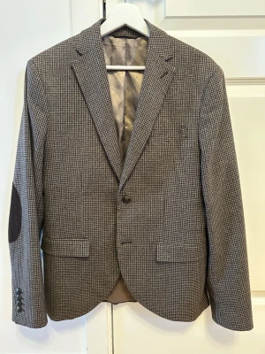 Grå rutig kavaj - Stilren grå kavaj med smått rutigt mönster och mörkbruna armbågslappar i mockaimitation. Tvåknäppt modell med klassisk krage, tre knappar vid ärmslut och två fickor framtill. Hel fodrad insida för extra komfort.