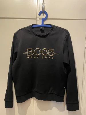 Svart sweatshirt från Hugo Boss - Svart sweatshirt med rund hals från Hugo Boss. Tröjan har ett stort guldfärgat BOSS-tryck på bröstet och ribbade muddar vid ärmslut och nederkant. Klassisk och stilren design som passar till det mesta. Liten i storleken.