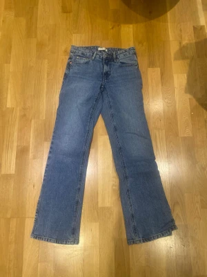Blå bootcut jeans med hög midja från Gina tricot - Säljer ett par blå bootcut jeans med hög midja och fem fickor. Jeansen har en klassisk blå tvätt och raka ben som slutar i en lätt utsvängd form. Tillverkade i denim, troligtvis bomull. Inga synliga loggor eller märken på bilderna.