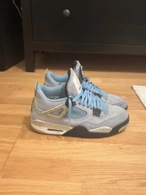 Jordan 4 University Blue  - Säljer ett par Air Jordan 4 University Blue sneakers i ljusblå mocka med meshpaneler och detaljer i svart och grått. Skorna har vita och grå inslag på sulan samt stänkdetaljer på hälen. Klassisk siluett med högt skaft och ikoniska AJ4-detaljer.
