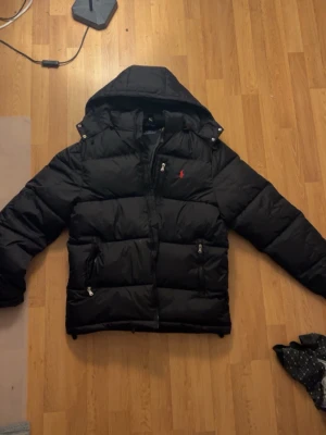 Svart dunjacka från Polo Ralph Lauren - Snygg svart dunjacka från Polo Ralph Lauren med huva och dragkedja framtill. Jackan har två stora fickor med dragkedja och en bröstficka. Klassisk röd logga på bröstet och quiltad design som ger en puffig look. Perfekt för kalla vinterdagar.