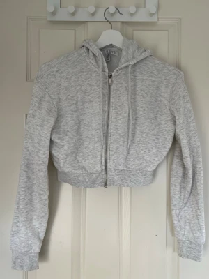 Ljusgrå cropped hoodie med dragkedja - Säljer en ljusgrå cropped hoodie/kofta från H&M med dragkedja framtill och huva med dragsko. Hoodien har långa ärmar och ribbade muddar vid ärmslut och nederkant. Perfekt till ett avslappnat streetwear-look.