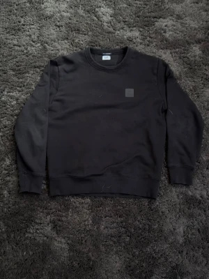 Svart sweatshirt från C.P. Company - Säljer en stilren svart sweatshirt från C.P. Company med rund halsringning och diskret logopatch på bröstet. Tröjan har ribbade muddar vid ärmslut och nederkant, perfekt för en clean och enkel look. Pris kan diskuteras.