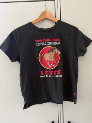 Svart Levi's t-shirt med cowboytryck - Cool svart t-shirt från Levi's med ett stort tryck på framsidan som visar en häst och texten 'Worn by all cowboys'. T-shirten har rund hals och korta ärmar. Perfekt för dig som gillar westernstil och vill sticka ut.