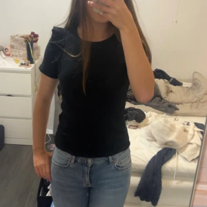 Svart ribbad kortärmad topp med volang - En enkel och stilren svart ribbad topp med korta ärmar och rund hals. Perfekt att matcha med jeans eller kjol för en clean look. Sitter figurnära och är ett måste i basgarderoben.