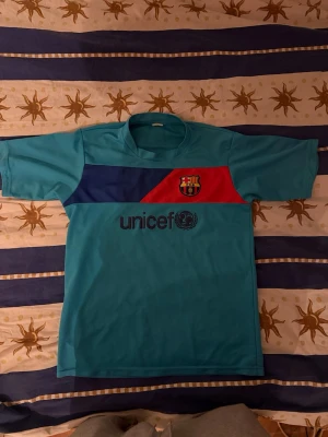 Turkos FC Barcelona Messi tröja - Säljer en turkos kortärmad matchtröja från FC Barcelona med Messi och nummer 10 på ryggen. Tröjan har klubbmärke på bröstet, UNICEF-logga framtill och färgblock i blått och rött. Perfekt för dig som älskar fotboll och vill stötta Barca.