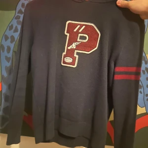 Mörkblå stickad tröja Polo Ralph Lauren - Mörkblå stickad tröja från Polo Ralph Lauren med stort rött P och vita detaljer på bröstet. Två röda ränder på ena ärmen ger en sportig vibe. Klassisk rund hals och ribbade muddar. Perfekt för dig som gillar college-stil.