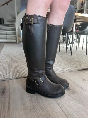 Mörkbruna höga Johnny Bulls läderstövlar med spännen - Säljer ett par mörkbruna höga Johnny Bulls stövlar i läder med dekorativa spännen vid ankeln och skaftet. Stövlarna har grov sula och dragkedja baktill för enkel på- och avtagning. Perfekta till jeans eller kjol för en cool look.