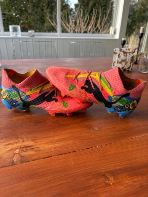 Färgglada Puma future fotbollsskor med brazil mönster - Säljer ett par riktigt unika fotbollsskor från Puma med färgstarkt mönster i rött, blått, gult och grönt. Skorna har hög strumpliknande krage, svart Puma-logga och coola detaljer med blommor och grafiska former. Perfekta för dig som vill sticka ut på planen!