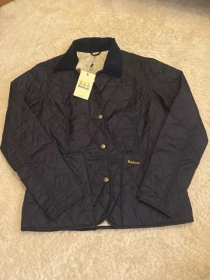 Mörkblå quiltad jacka från Barbour - Quiltad mörkblå jacka från Barbour med klassisk krage och tryckknappar framtill. Jackan har två framfickor och diskret Barbour-logga på ena fickan. Perfekt för kyliga dagar och har en tidlös design.