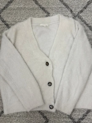 Vit fluffig kofta med knappar - Mysig vit kofta med fluffig känsla och tre stora knappar framtill. Koftan har v-ringning och långa ärmar. Tryck inte på Köp innan ni kontaktat mig!