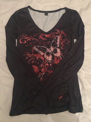Svart långärmad topp med fjärilstryck - Cool svart långärmad topp med v-ringning och stort rött tryck. Jättesnygg men säljer för att jag ångrade mig när tröjan kom. Materialet är lite skumt då det känns nästan som en träningströja. Aldrig använd