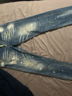 Dsquared2 blå jeans med färgstänk - Säljer ett par blå Dsquared2 jeans med coola slitningar och färgstänk i vitt och gult. Jeansen har smal passform, klassiska fem fickor och en gul etikett med texten 'Caten Heated' bak i linningen. Perfekta för dig som gillar streetwear och unika detaljer.