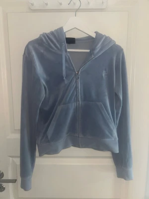 Blå velour hoodie med dragkedja - Snygg blå hoodie i mjuk velour med dragkedja framtill. Har huva med dragsko och långa ärmar. Perfekt för en chill och avslappnad stil. Liten broderad logga på bröstet ger extra detalj. Använt ett få tal gånge 