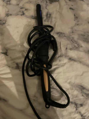 Svart locktång med beige detalj - En svart locktång med beige detalj på handtaget. Locktången har en sladd som är virad runt handtaget och ett smalt värmeelement. Perfekt för att styla håret och skapa lockar.