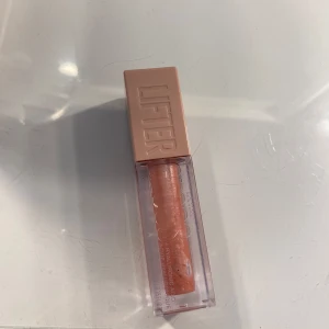 Läppglans med rosa nyans - Läppglans i en ljusrosa nyans med en fyrkantig, genomskinlig förpackning och ett beige lock. Perfekt för att ge läpparna en glansig finish. Färgen ”003, moon” 