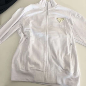 Prada hoodie (Vit) - Säljer en stilren vit hoodie från Prada med dragkedja och hög krage. Jackan har en diskret logga på bröstet och långa ärmar. Perfekt för en minimalistisk look.