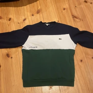 Lacoste sweatshirt  - Snygg tröja från Lacoste i tre färger: mörkblå, vit och grön. Den har en klassisk design med Lacoste-loggan broderad på bröstet och texten 'Lacoste' tryckt på den vita delen. Perfekt för en stilren och avslappnad look. Strl medium passar en medium