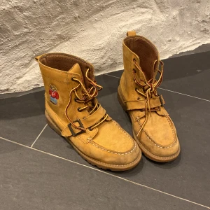 Polo Ralph Lauren Bruna boots - Snygga bruna boots i skinn från Polo med ett broderat björnmotiv på sidan. Skorna har snörning och en robust sula, perfekt för höst och vinter. De har en klassisk design med en modern twist tack vare det charmiga motivet. Storlek 34,5