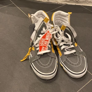 Grå och gula Vans sneakers med dragkedja - Snygga Vans sneakers i grått och gult med dragkedja och snörning. Skorna har en klassisk vit sula och den ikoniska Vans-loggan på sidan. Perfekta för en cool och avslappnad stil.